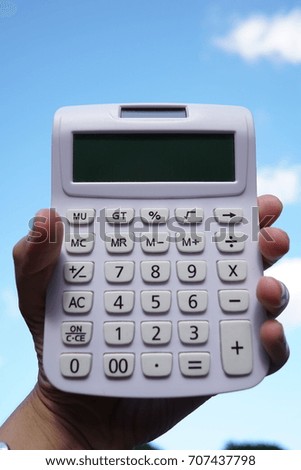 A man's hand holding a white calculator with blue sky and white cloud background.