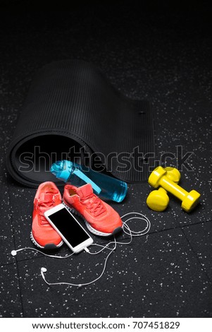 A close-up picture of a blue bottle for water, black stretching mat, bright pink training shoes, yellow equipment for physical exercises and modern phone with handphones on a spotted black background.