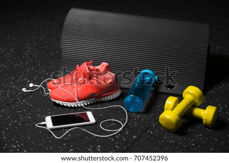 A close-up picture of bright pink sports shoes, yellow dumbbells, blue sportive bottle and black pilates mat on a black background. A colorful composition of sportive accessories. Healthy lifestyle.