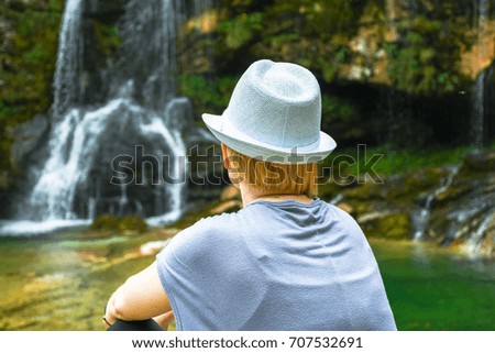 Woman with blond hair and white hat looking, sitting and relaxing by the natural small lake and waterfall in the background 
