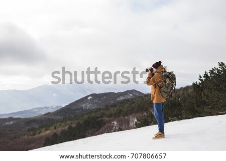 Tourist taking photo of winter mountains. Outdoor style, beige parka, backpack, jeans