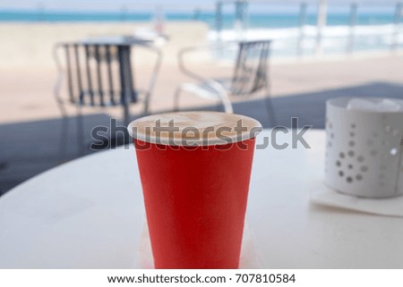 Red disposable cup of coffee standing on a white table on blue sea and beach background