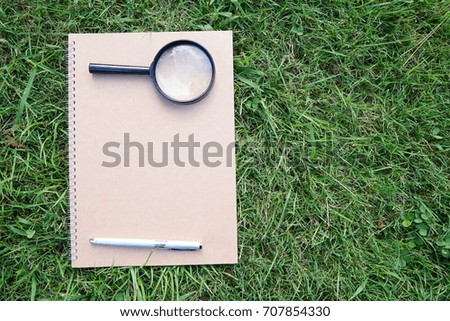 Sketchbook with magnifier or magnifying glass and ball pen on the green grass.
