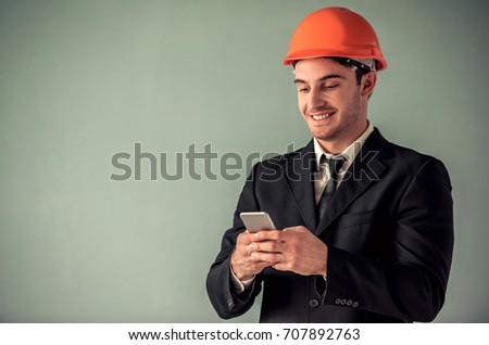 Handsome young architect in suit and protective helmet is using a smart phone and smiling, on gray background