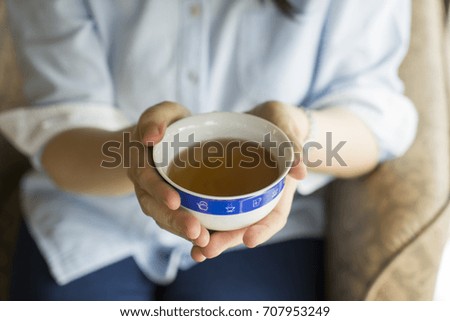 Woman holding white ceramic cup of hot Chinese tea with both hand, sitting on a chair in living room.