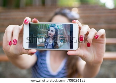 Closeup shot of a young attractive female wearing casual clothes taking selfie by holding smart phone with both hands while enjoying outside on a sunny day. Putting her lovely mood on the line