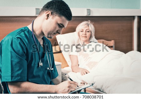 Handsome young doctor in uniform with phonendoscope on his neck writing down complaints of patient, who is lying in the hospital bed in the hospital ward. Healthcare concept