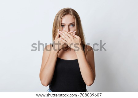 Blonde woman with shining eyes, covering her mouth with hands while trying to keep silence and not telling secret of her best friend. Young female model making hush sign, isoated over white wall