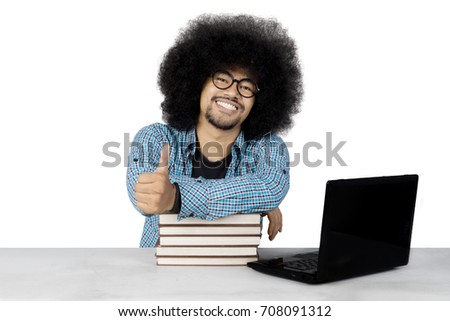 Photo of an Afro male college student showing thumb up while studying with a laptop and books on the desk