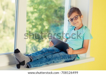 Little boy sits on the windowsill and listening to music through headphones. Child uses the tablet, watching cartoon, playing games, surfing internet.