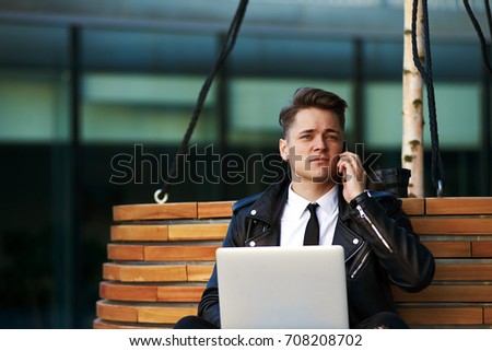 Always in touch. Picture of attractive young Caucasian male entrepreneur sitting on bench with laptop pc, using free wi-fi for remote work, checking e-mail and talking on mobile phone simultaneously.