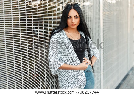 Fashionable beautiful model of a woman in a white cape with patterns near the wall of a grid