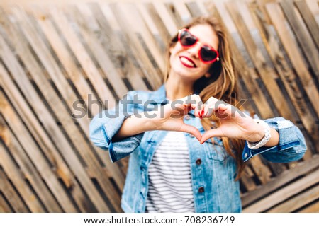 Portrait of young european woman in glasses making heart sign to express her love feelings. Charming girl in denim suit posing making love sign on the blur background. Focus on hands, body part