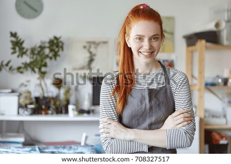 Waist up shot of cheerful young Caucasian female creative worker, designer or artist with long ginger hair and freckles having inspired expression on her face while working in her modern workshop