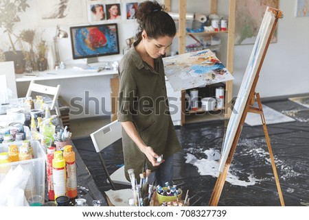 Creative female making strokes of brush on easel standing in her workshop, surrounded with different colorful oils. Talented painter drawing picture in art studio using watercolors and paint brush.