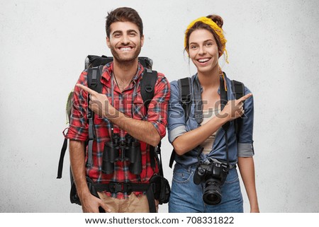Studio shot of attractive adventurous young couple in love wearing practical clothes, carrying rucksacks, photo camera and binoculars having cheerful looks, pointing fingers in opposite directions