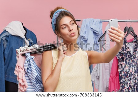 Pretty young female making selfie while standing near racks with clothes, being glad to spend her free time in shopping centre. Adorable lady using modern mobile phone while doing shopping alone.
