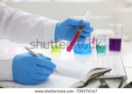 Employee of the chemical laboratory teacher chemist holds a silvery pen in his hand makes notes in the diary records test data from reactions examining test tubes with the substance arm in gloves Royalty-Free Stock Photo #708376567