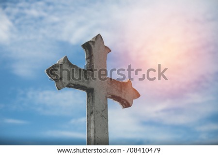 Stone cross with sun background against blue sky and clouds. Conceptual stone cross religion symbol.