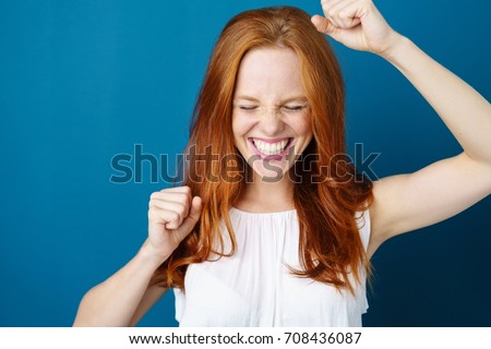 Attractive young redhead woman celebrating a victory punching the air with her fists and a beaming toothy smile over a blue studio background with copy space Royalty-Free Stock Photo #708436087