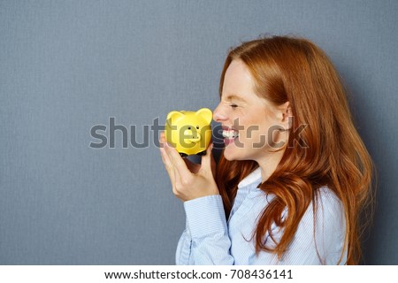 Young red-haired woman kissing yellow piggy bank against blue background
