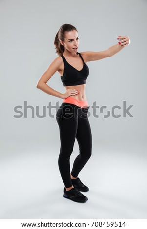 Full length picture of pretty fitness woman posing with arm on hip and making selfie on her smartphone over gray background