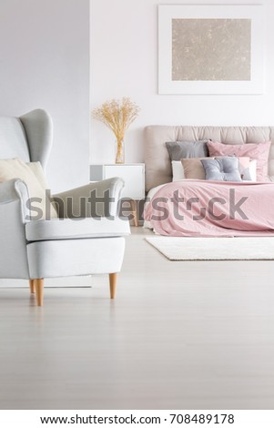 Grey armchair with pillow in trendy bedroom with flowers on white bedside cabinet 