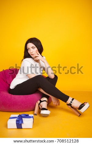  Girl sitting on a pink cushioned chair on a bright yellow background.Dressed in a white shirt and blue Trouser suit and black sandals with high heels,business style.In the hands holding the gift