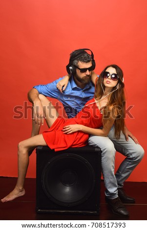 Technologies and music concept. Couple in love lies on black loudspeaker on red background. Guy with beard and lady hug and listen to music. Man with beard wears headphones and sunglasses.