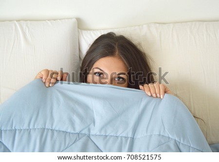 Beautiful lazy young woman lying down in the bed and sleeping. Teen girl with open eyes covers her face with blanket in the morning. Do not get enough sleep concept. Copy space.