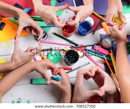 Artists hands with stationery and colored paper. Messy art concept. Hands hold colorful markers, pencils and paints. Art supplies in hands showing heart signs on white wooden desk background, top view