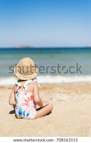 Little girl child with straw hat and floral swimsuit looking at the sea in a sunny day. Little girl sitting back on the shore of the sea