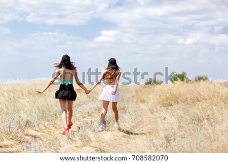 A full-length photo of couple girls wearing colorful clothes on a natural blue sky background. Two glamorous young ladies walking hand in hand along a field and enjoying a hot summer. Copy space.