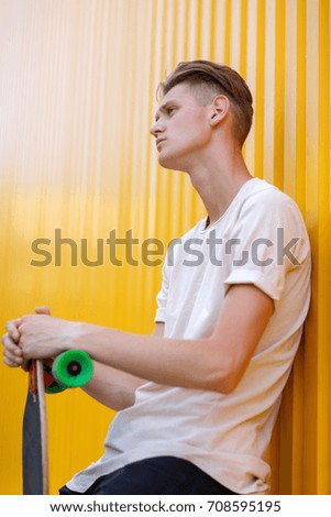 A close-up photo of a fashion young man wearing casual clothes with a modern skateboard on a bright yellow wall background. Motion, skateboarding, sports, action, and training concept. Copy space.