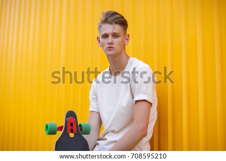 A close-up photo of a fashion young man wearing casual clothes with a modern skateboard on a bright yellow wall background. Motion, skateboarding, sports, action, and training concept. Copy space.