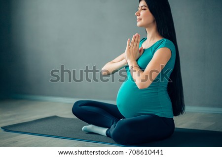 Portrait of a beautiful young pregnant woman holding her stomach while sitting near an exercise ball in the sport center. Working out, yoga and fitness, pregnancy concept.