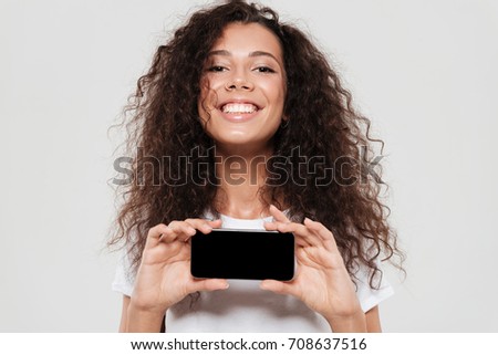 Close up picture of smiling curly woman showing blank smartphone screen and looking at the camera over gray background