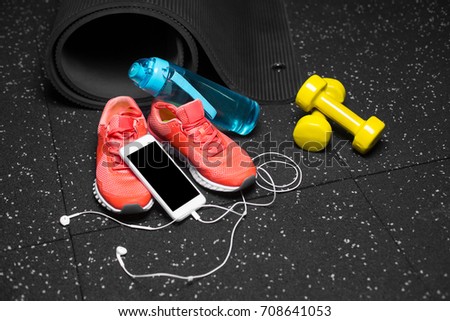A close-up picture of a sports shaker for water, black stretching mat, bright pink training shoes, yellow equipment for physical exercises and new phone with handphones on a spotted black background.