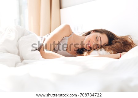 Image of young smiling pretty lady lies in bed indoors. Eyes closed.