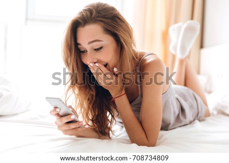Image of young happy emotional pretty lady lies on bed indoors. Looking aside chatting by phone.