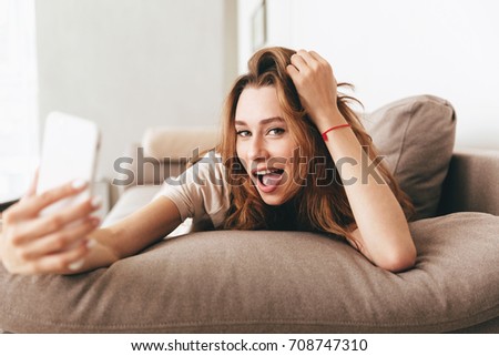 Image of young amazing emotional pretty lady lies on sofa indoors. Looking camera make selfie by phone.