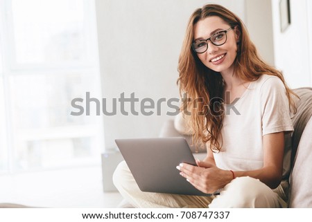Image of young happy amazing pretty lady sitting on sofa indoors. Looking camera using laptop computer.