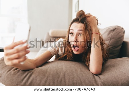 Image of young shocked emotional pretty lady lies on sofa indoors. Looking aside make selfie by phone.