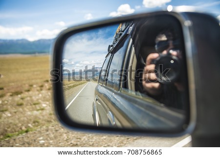 Man or woman taking photo with professional camera in a car side mirror with road and natural mountain background. asphalt road in perspective. blue sky with clouds