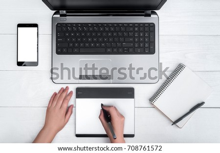 Top view of open laptop computer, cell phone, empty diary and graphic tablet. Girl's hands with graphic tablet drawing and retouching image on laptop, free space. Mobile phone with white screen