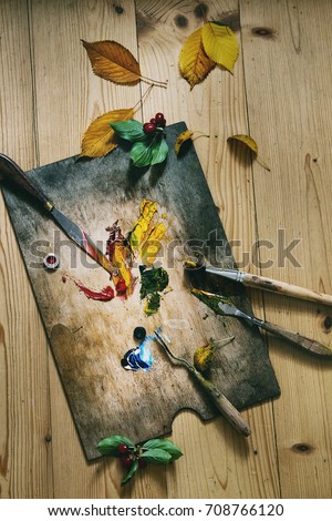 Wooden palette with oil paints yellow red, brushes and palette knife over wooden floor. With autumn decoration yellow leaves and red berries. Top view with space. Art concept. Atmospheric day light.