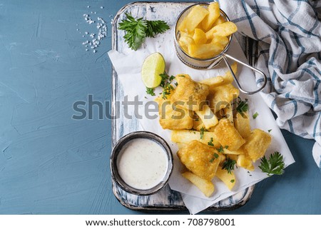 Traditional british fast food fish and chips. Served with white cheese sauce, lime, parsley, french fries in frying basket on white paper over blue concrete background. Top view, copy space.