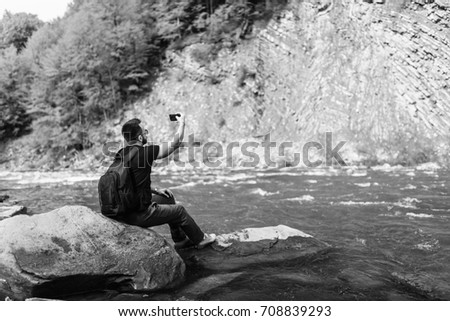 Traveler with backpack making photo on mobile phone near mountain river, black and white