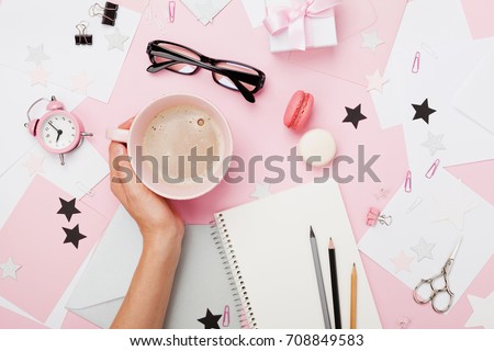 Female hand with coffee cup, macaron, office supply, gift and notebook on pastel desk top view. Fashion pink woman workplace background for blogging. Flat lay. Beautiful morning breakfast.
