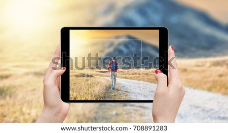 Female hands holding tablet and taking photo of man with backpack on the top of the mountain in sunset